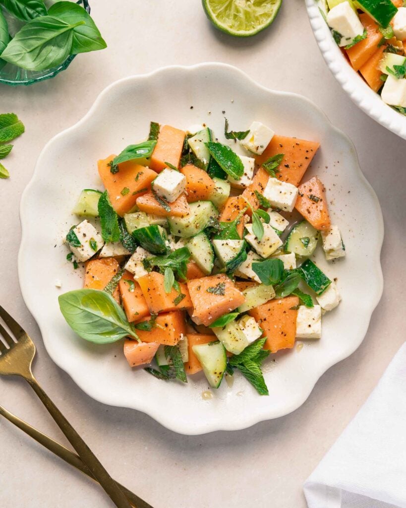 Une assiette blanche avec une salade de melon, de concombre et de feta garnie de feuilles de menthe fraîche et de basilic, servie à côté d'ustensiles dorés et d'un citron vert coupé.