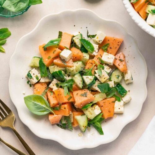 Une assiette blanche avec une salade de melon, de concombre, de feta, de menthe fraîche et de feuilles de basilic, garnie de poivre noir. Une fourchette et un couteau sont placés à côté de l’assiette, avec un citron vert tranché à proximité.