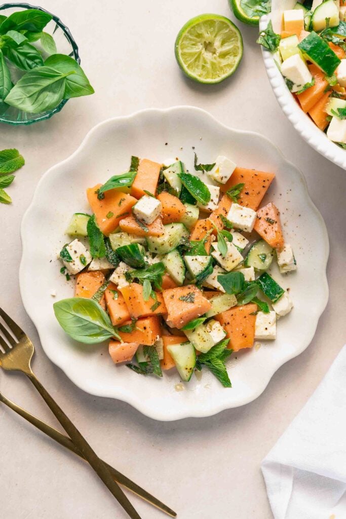 A white plate with a salad of melon, cucumber, feta, fresh mint and basil leaves, garnished with black pepper. A fork and knife are placed beside the plate, with a sliced lime nearby. 