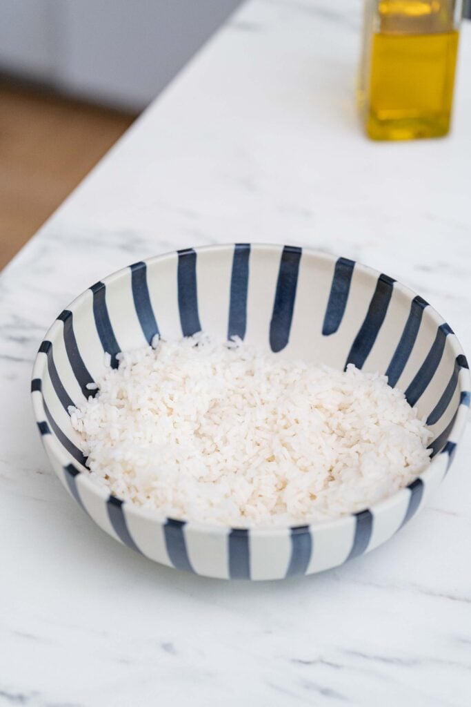 Un bol rayé bleu et blanc rempli de riz blanc repose sur un comptoir en marbre, avec une bouteille d'huile d'olive visible en arrière-plan.