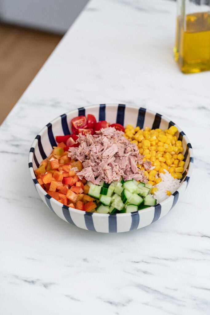 A striped bowl on a marble countertop holds rice, tomatoes, corn, rice, cucumbers, peppers and crumbled tuna.