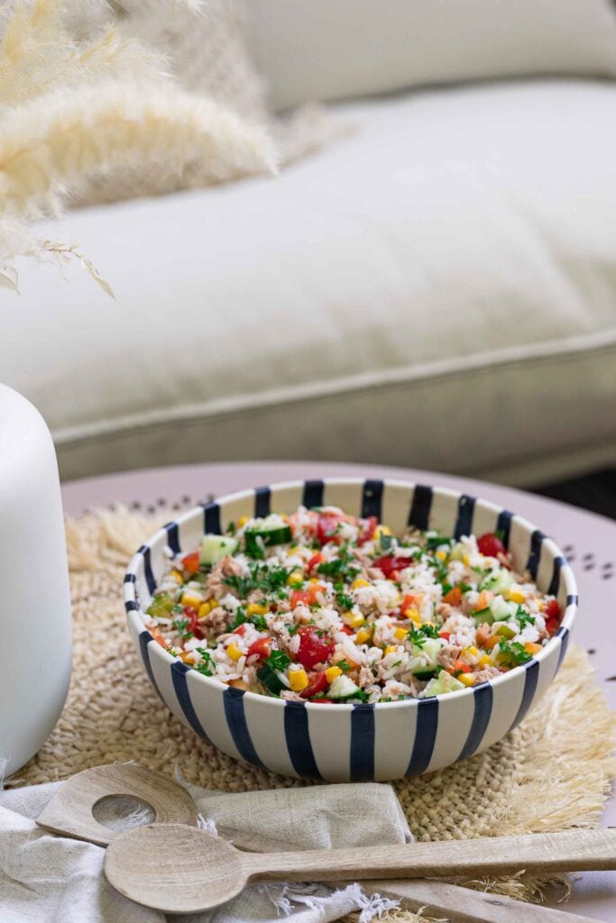 Un bol de salade de riz composée de légumes posé sur un set de table texturé, à côté de cuillères en bois et d'un vase blanc, avec un canapé en arrière-plan.