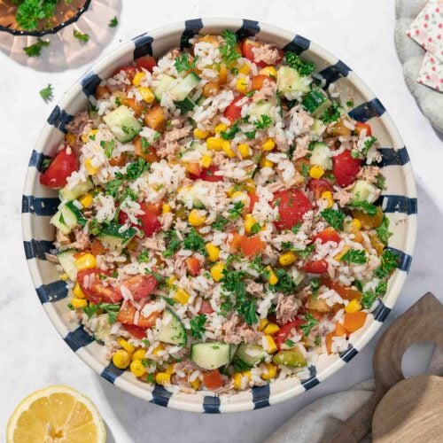 Un grand bol rayé rempli d'une salade de riz colorée composée de tomates, de concombres, de maïs et de divers légumes verts. Deux ustensiles en bois et des tranches de citron sont placés à proximité sur une surface claire.