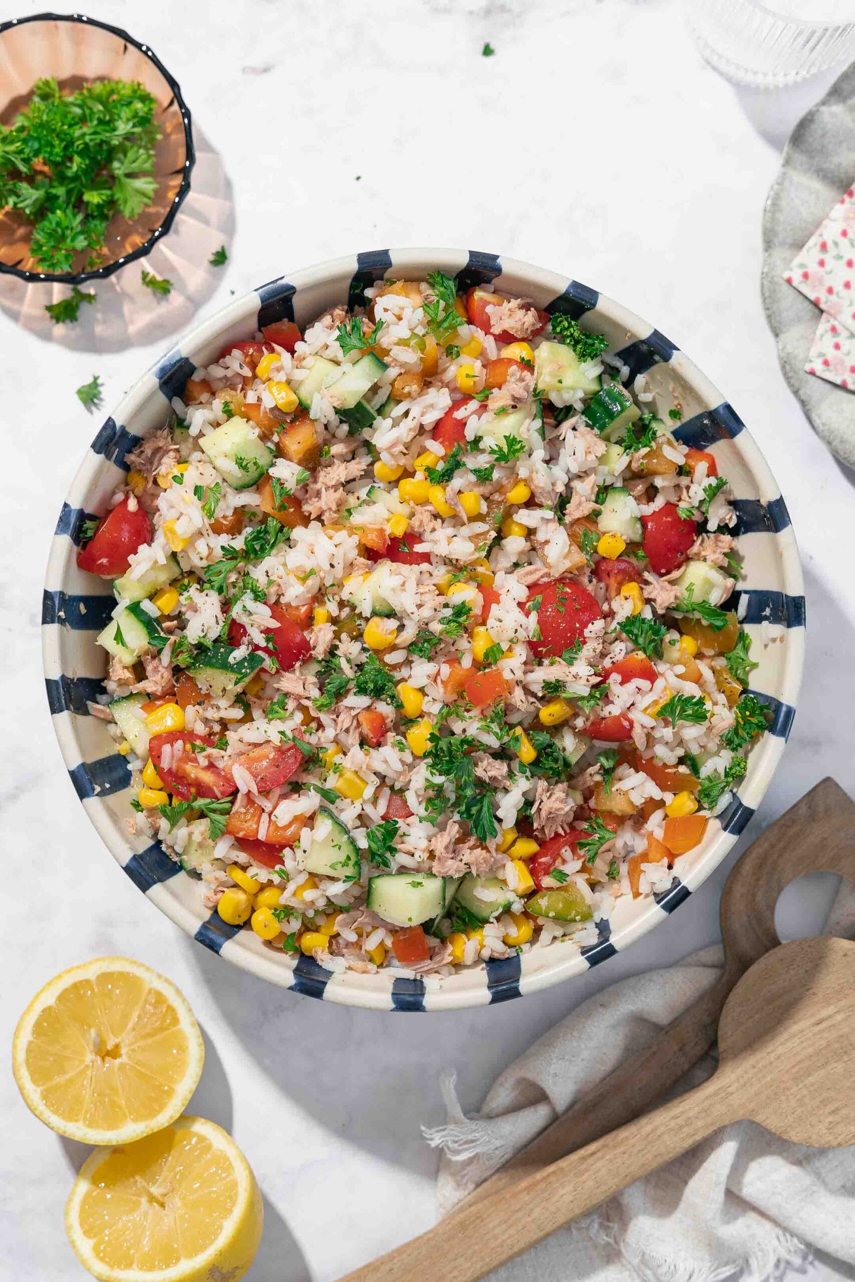 Eine große, gestreifte Schüssel, die mit einem bunten Reissalat aus Tomaten, Gurken, Mais und verschiedenen grünen Gemüsesorten gefüllt ist. Zwei hölzerne Utensilien und Zitronenscheiben werden in der Nähe auf einer hellen Oberfläche platziert. 