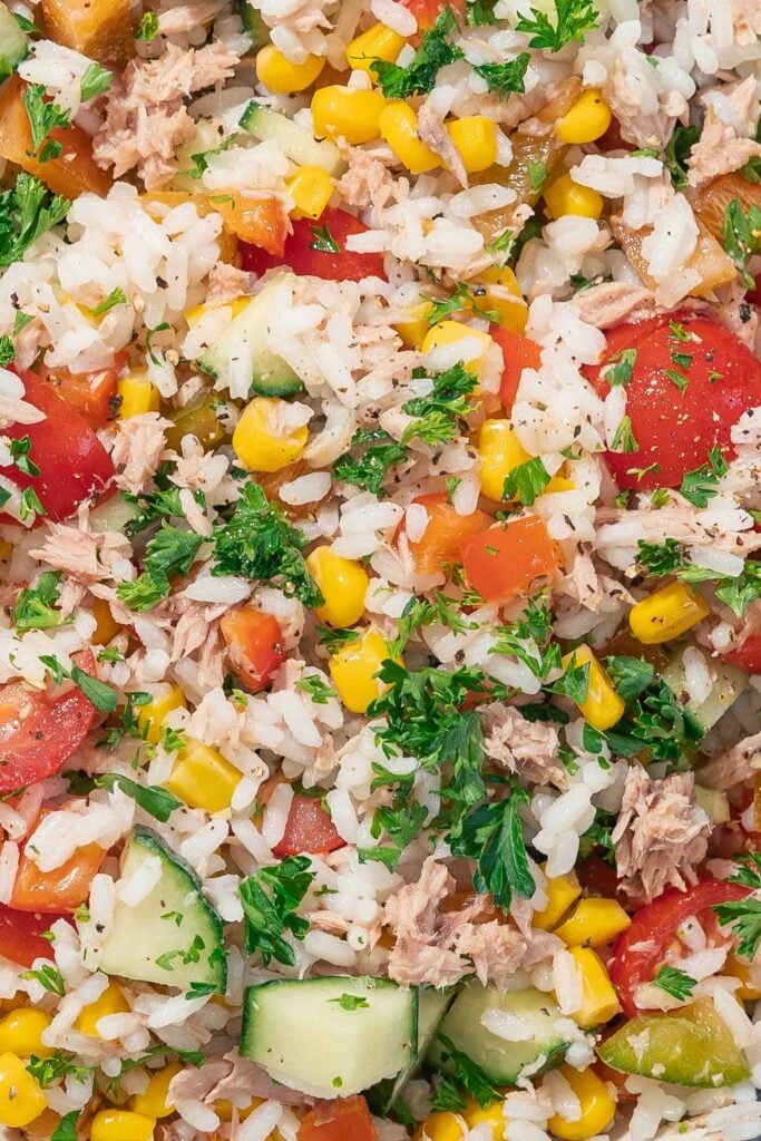 Close-up of a mixed rice salad containing white rice, diced tomatoes, corn, cucumber pieces, grated tuna and chopped parsley.