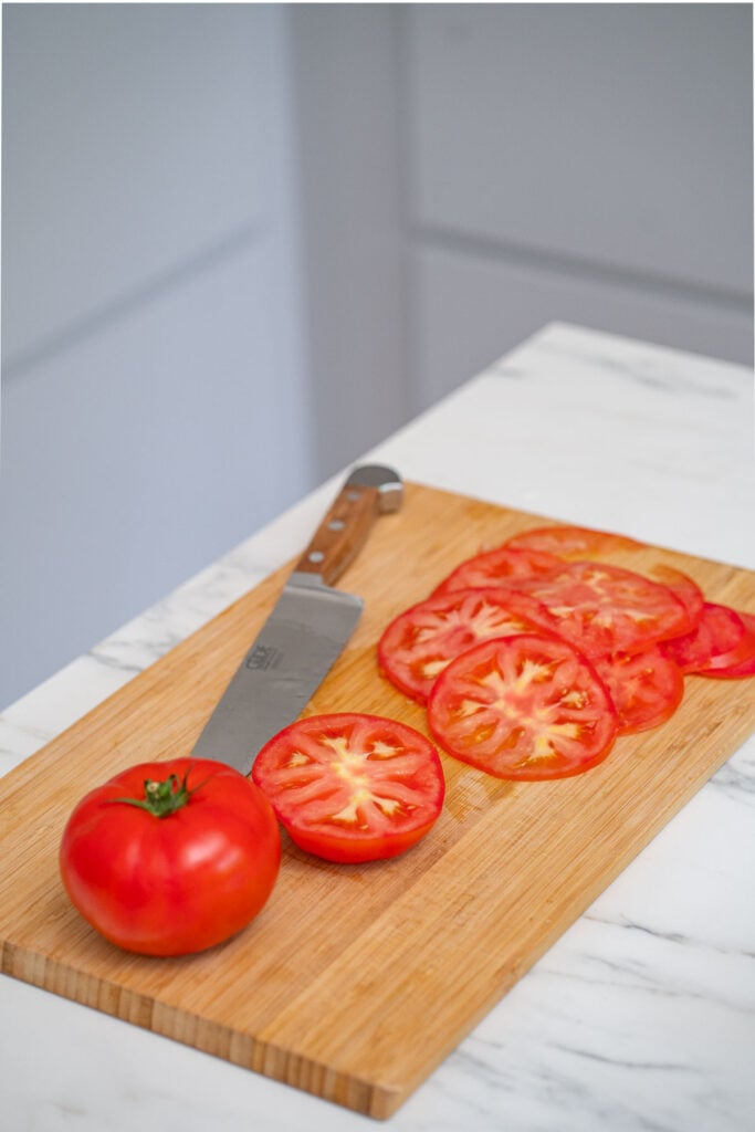 A cutting board with a knife, a whole tomato and thin slices of tomato.