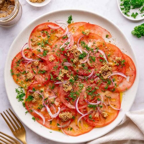 Une assiette blanche avec des tomates finement coupées, des oignons rouges, des herbes hachées et des graines de moutarde. Un pot de graines de moutarde, un petit bol d'herbes et une serviette en tissu avec des fourchettes dorées se trouvent à proximité.