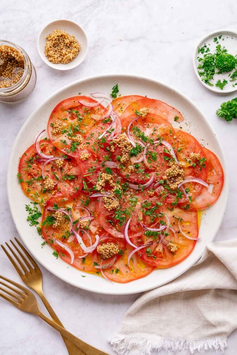 Une assiette blanche avec des tomates finement coupées, des oignons rouges, des herbes hachées et des graines de moutarde. Un pot de graines de moutarde, un petit bol d'herbes et une serviette en tissu avec des fourchettes dorées se trouvent à proximité.