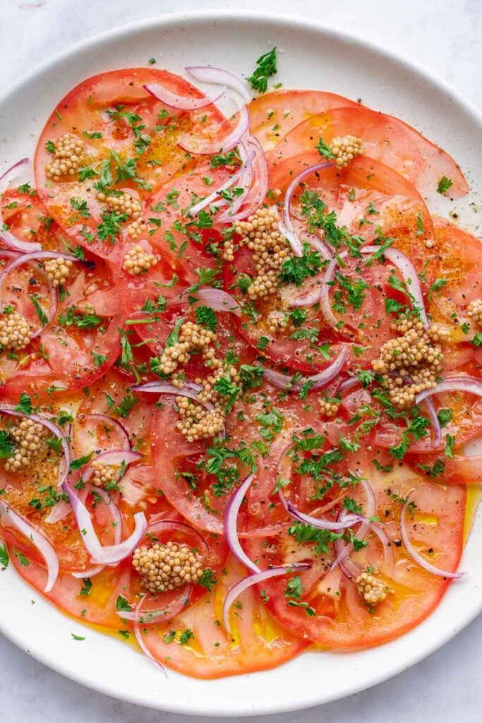 A plate of sliced tomatoes topped with thinly sliced red onions, whole mustard seeds, chopped parsley and seasonings.