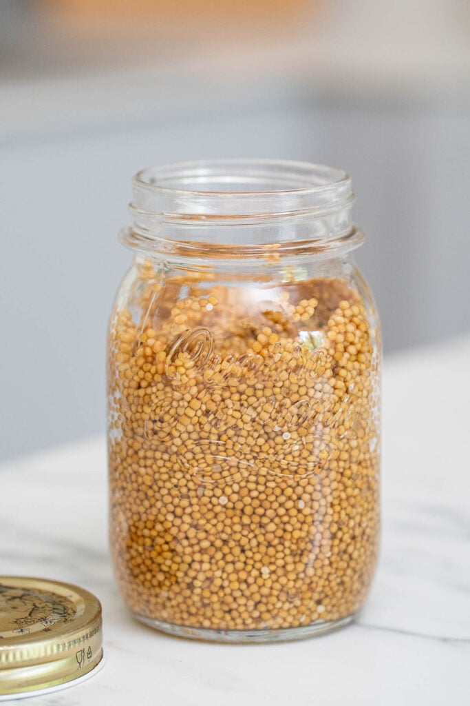 A clear glass jar filled with em pickles yellow mustard seeds, with the metal lid placed on a marble countertop next to it.