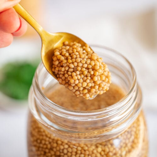 A hand with red nail polish holding a gold spoon retrieves pickled mustard seeds from a jar filled with mustard seeds.