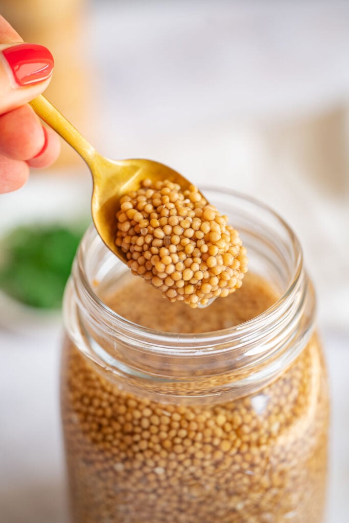 Eine Hand mit rotem Nagellack, die einen goldenen Löffel hält, holt Senfkaviar aus einem mit Senfkörnern gefüllten Glas.