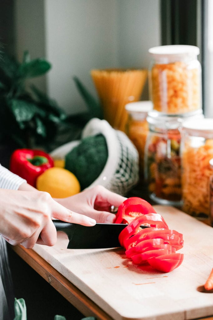 Personne coupant des tomates sur une planche à découper en bois dans une cuisine avec des pots de pâtes et de légumes en arrière-plan.