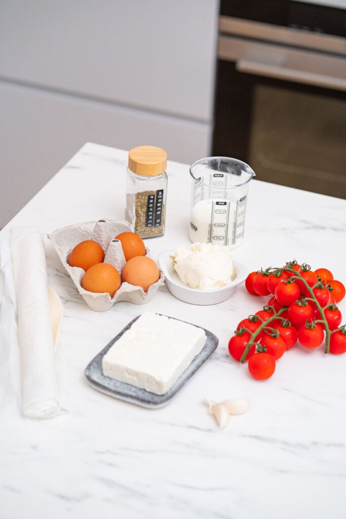 Un comptoir avec des ingrédients de cuisine comprenant des œufs, du poivre noir, de l'huile d'olive dans une tasse à mesurer, des tomates cerises, de l'ail, un bloc de fromage sur une assiette, du fromage à la crème et un rouleau de pâte pour préparer une quiche aux tomates cerises et feta.