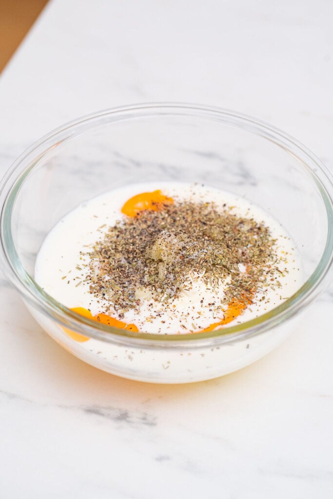 A clear glass bowl containing a mixture of eggs, milk and various spices on a white marble surface.