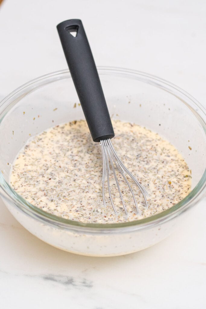 A glass bowl containing a creamy mixture of herbs and spices, with a black-handled whisk resting inside.