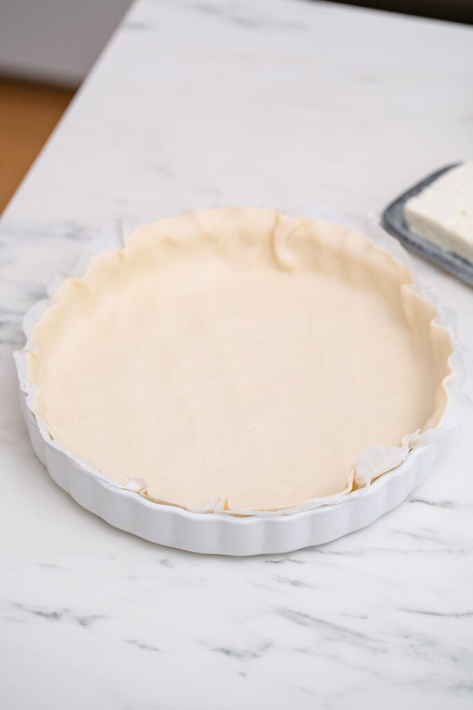 A white baking dish with an unbaked pie crust on a marble countertop, with a block of butter in the background.