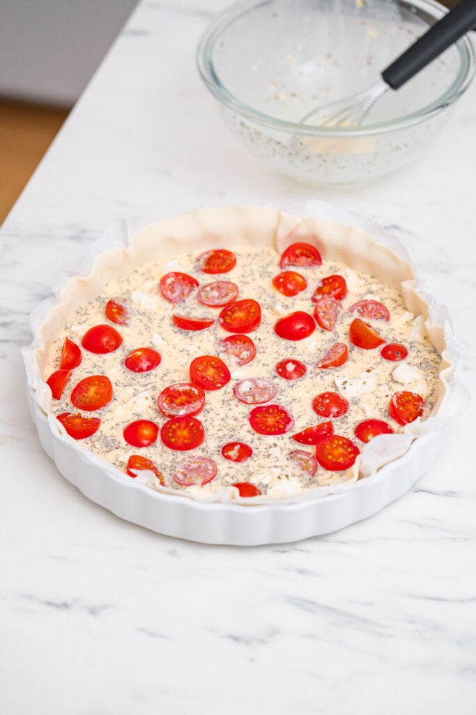 Une croûte à tarte ronde remplie d'un mélange et garnie de tomates cerises tranchées sur une surface en marbre blanc ; un fouet et un bol en verre sont à l'arrière-plan.