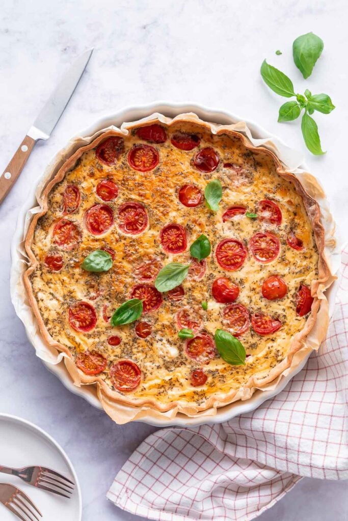 Eine Quiche mit Kirschtomaten und Feta, die auf einer Tortenplatte auf einer Marmorfläche platziert ist. Ein Messer, eine Stoffserviette und ein Basilikumzweig liegen daneben. 