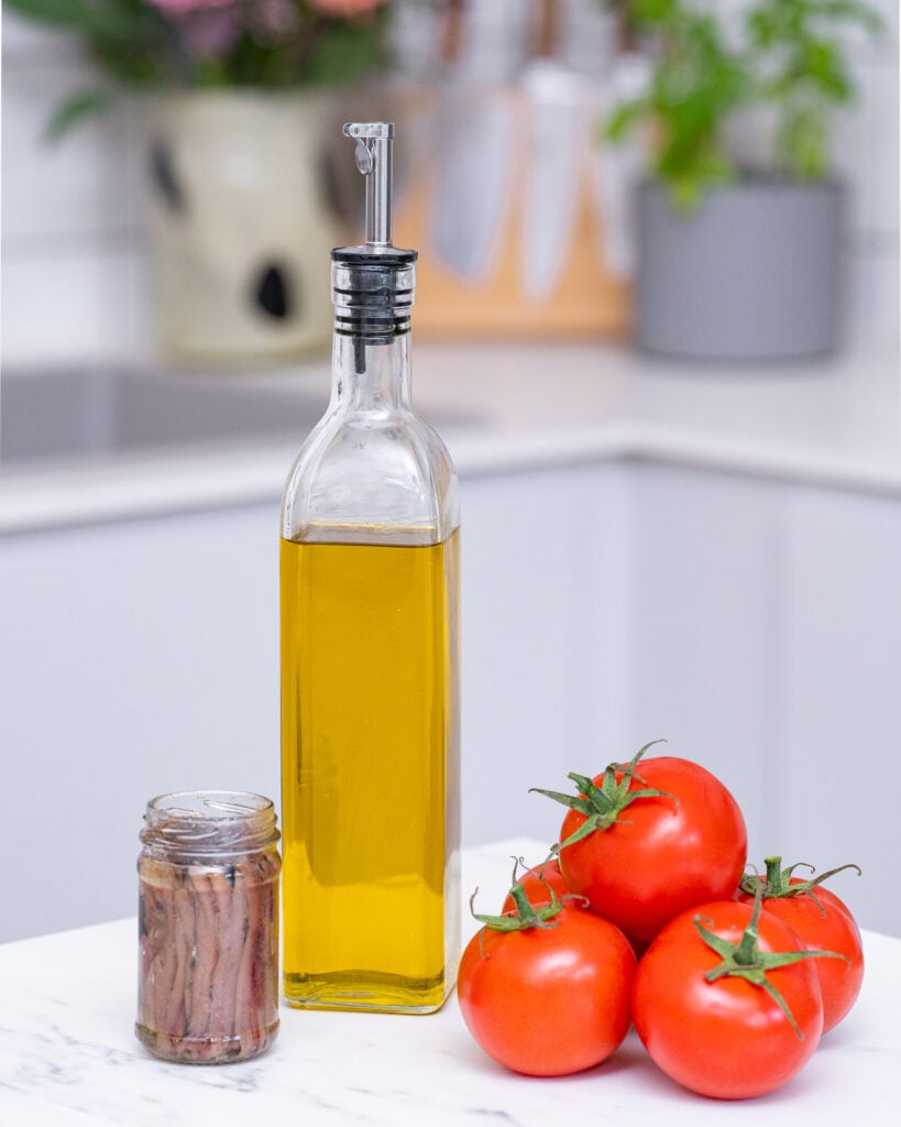 A bottle of olive oil, a jar of anchovies and three ripe tomatoes lie on a kitchen worktop.