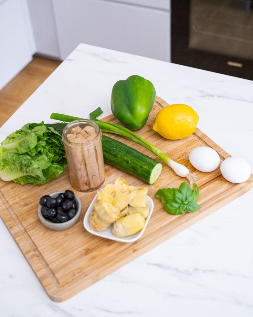 A wooden cutting board on a kitchen counter with lettuce, black olives, artichoke hearts, tuna in a jar, green bell pepper, cucumber, lemon, green onions, basil leaves and two hard-boiled eggs.