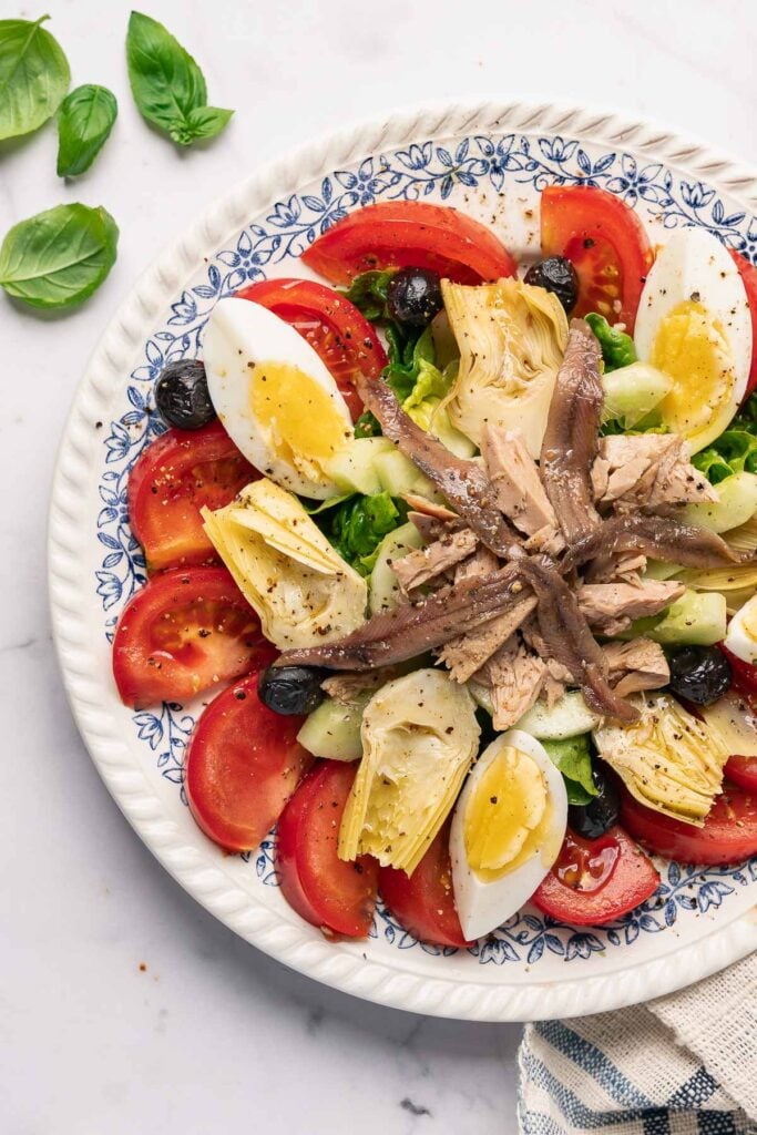 Une vraie salade Niçoise sur une assiette décorative comprend des tomates, des œufs durs, des anchois, du thon, des olives, des artichauts et des légumes-feuilles. Des feuilles de basilic et un tissu rayé sont en arrière-plan.