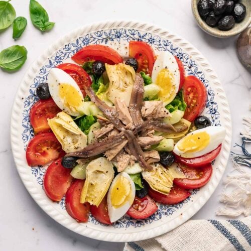 La vraie recette de la salade niçoise avec des tranches de tomates, des œufs durs, des olives noires, des cœurs d'artichauts, des anchois et du thon. Des feuilles de basilic frais et un petit bol d'olives sont à proximité.