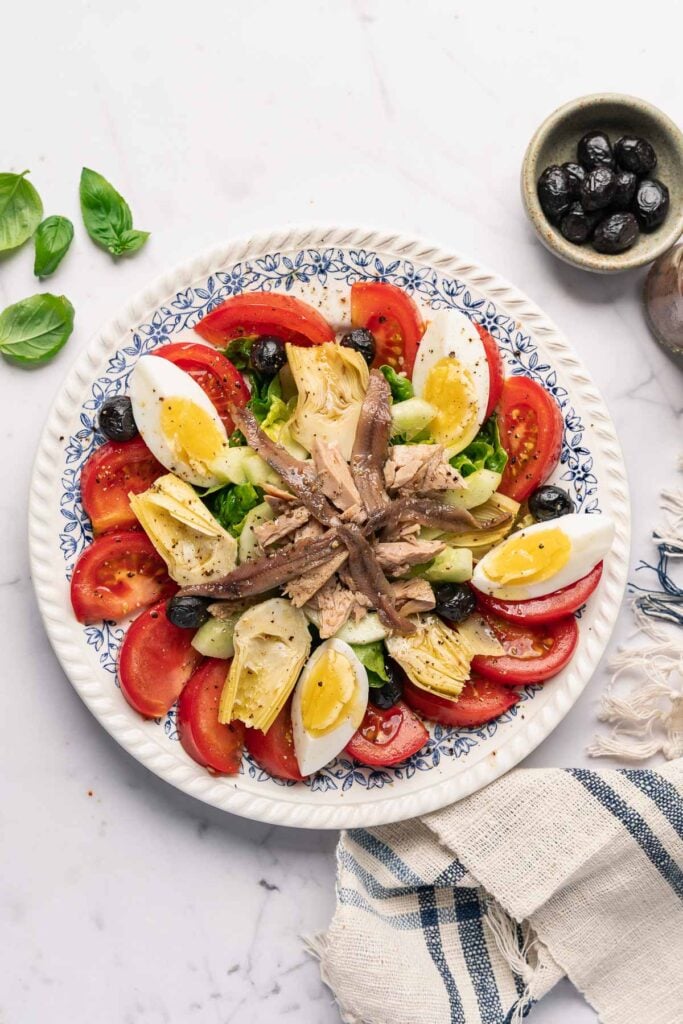 La vraie recette de la salade niçoise avec des tranches de tomates, des œufs durs, des olives noires, des cœurs d'artichauts, des anchois et du thon. Des feuilles de basilic frais et un petit bol d'olives sont à proximité.