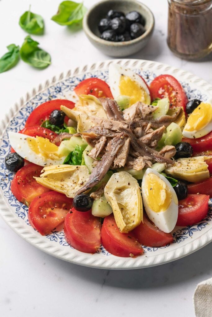 Une vraie salade Niçoise sur une assiette composée de quartiers de tomates, d'œufs durs, de concombres, d'olives noires, de cœurs d'artichauts et de filets d'anchois, garnie d'herbes et d'un bol d'olives en arrière-plan.