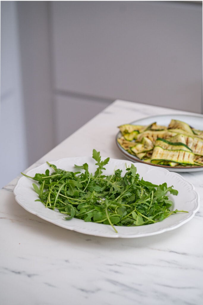 Une assiette blanche avec de la roquette fraîche sur un comptoir en marbre se trouve à côté d'un plat séparé comportant une salade de courgettes grillées en arrière-plan.