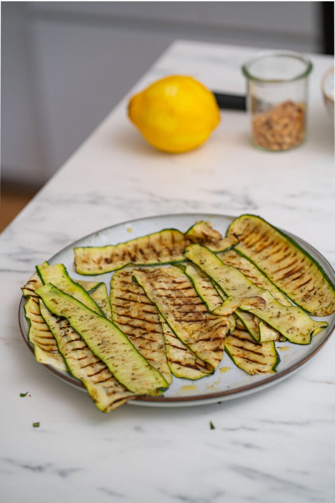 Slices of grilled zucchini arranged on a plate on a marble countertop, with a lemon and a glass jar in the background.