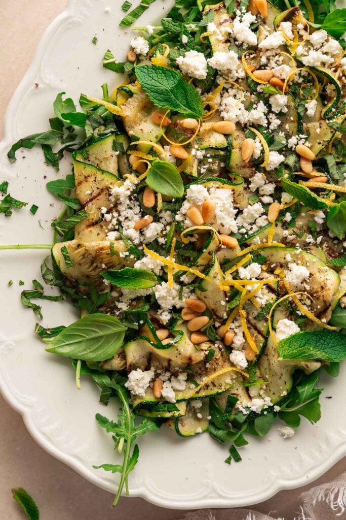 Une assiette blanche de tranches de courgettes grillées garnies de fromage feta émietté, de pignons de pin, de zeste de citron et de feuilles de basilic et de menthe fraîches, servies sur un lit de légumes verts à feuilles.