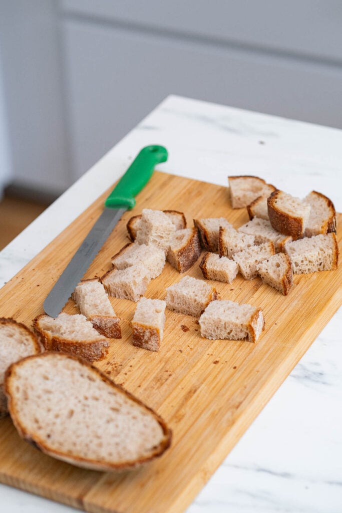 Schneiden Sie Scheiben und Würfel von Sauerteigbrot auf einem hölzernen Schneidebrett mit einem gezackten Messer mit grünem Griff.