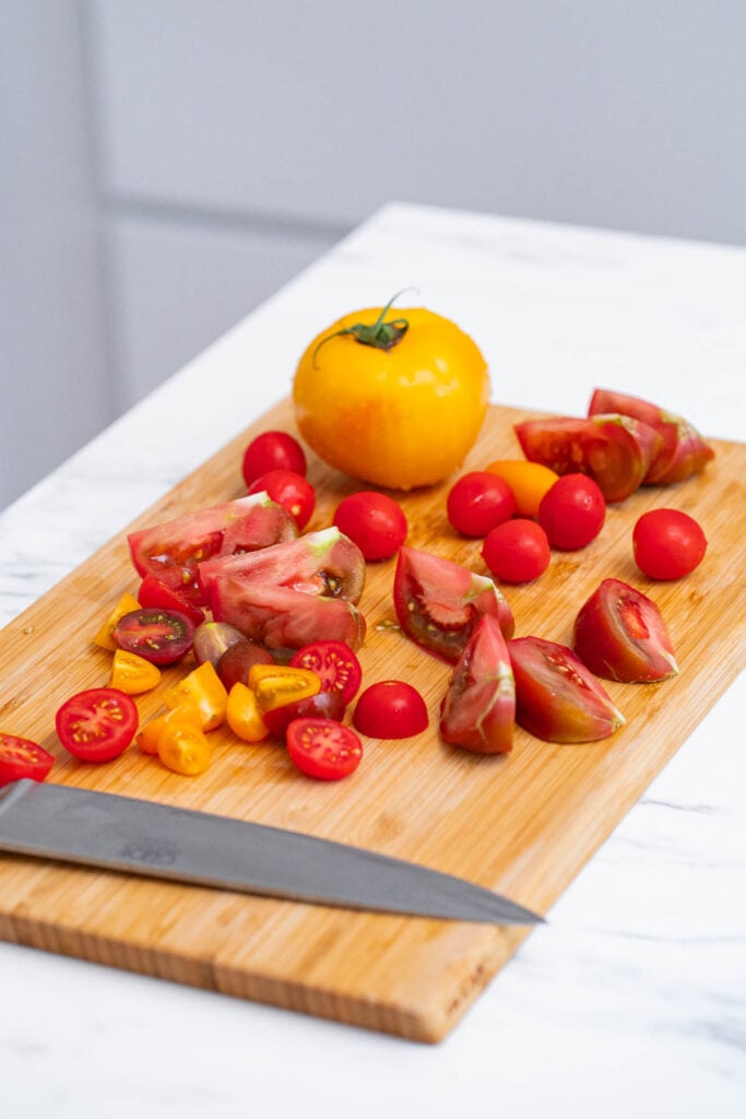 A chopping board on a white countertop with a variety of chopped tomatoes, including red, yellow and green heirloom tomatoes, and a knife.