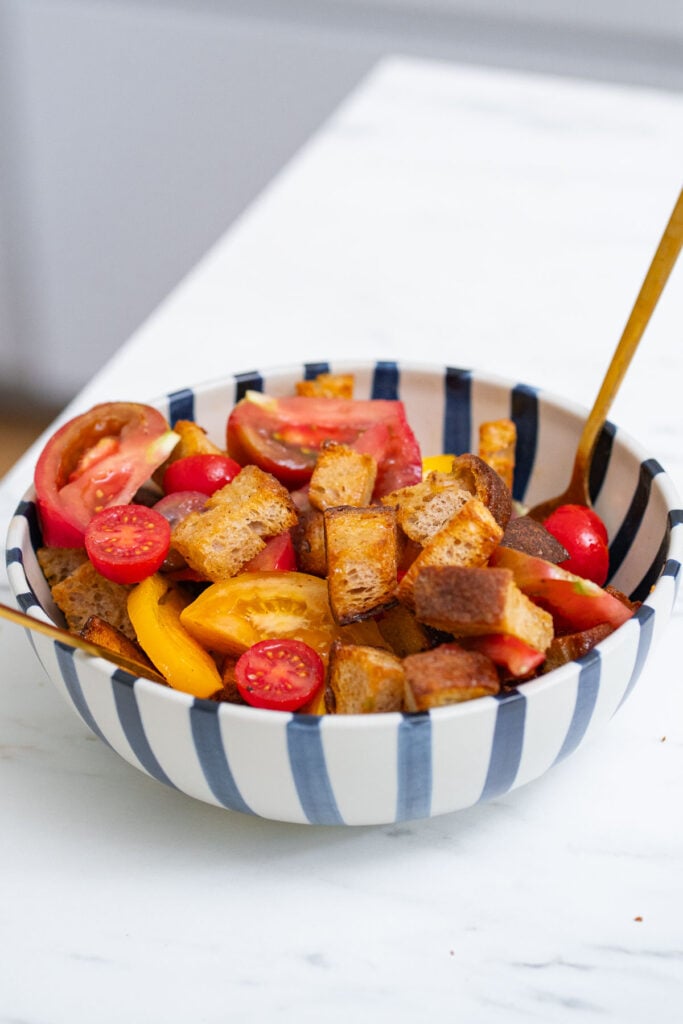 A bowl containing croutons, sliced tomatoes, cherry tomatoes and yellow peppers with a fork on a white marble surface.