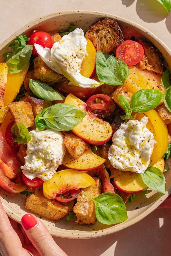 Un bol de salade composé de légumes verts mélangés, de tranches de pêches, de tomates cerises, de croûtons, de feuilles de basilic frais et de cuillerées de fromage burrata, avec une main tenant le bord du bol.