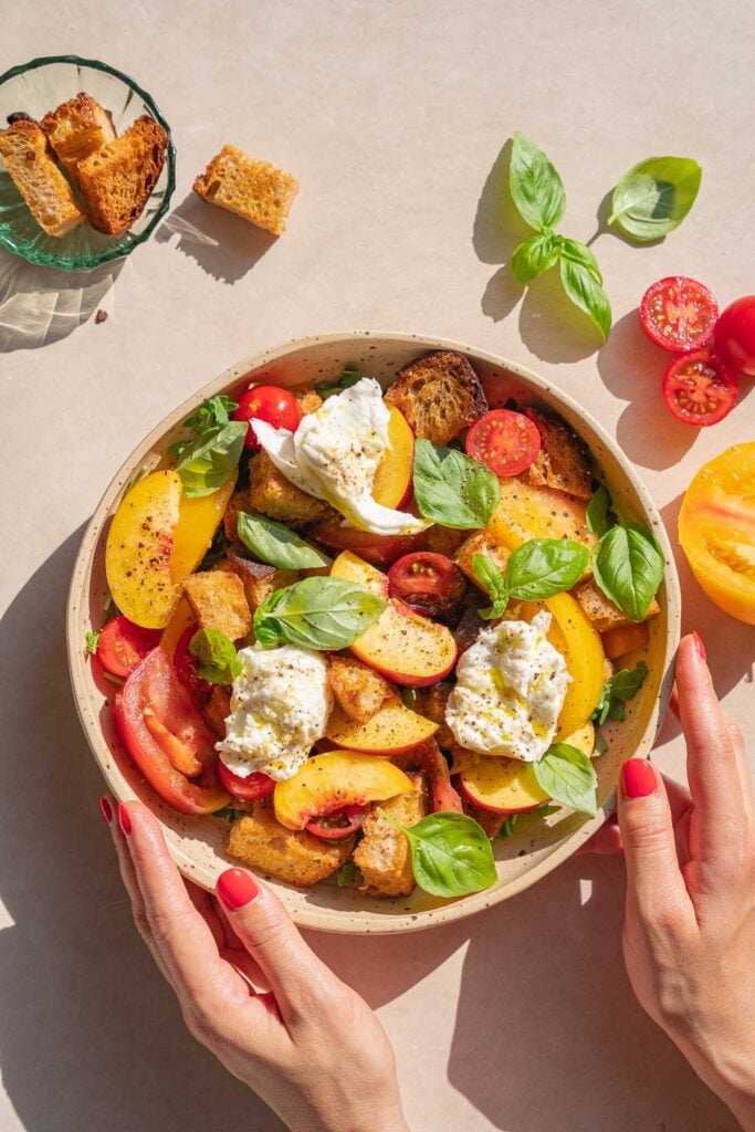 Une personne tient un bol de panzanella à la pêche et burrata contenant des tomates, des feuilles de basilic, des croûtons et des morceaux de fromage crémeux. Autour du bol se trouvent d'autres croûtons, des tomates et des feuilles de basilic.