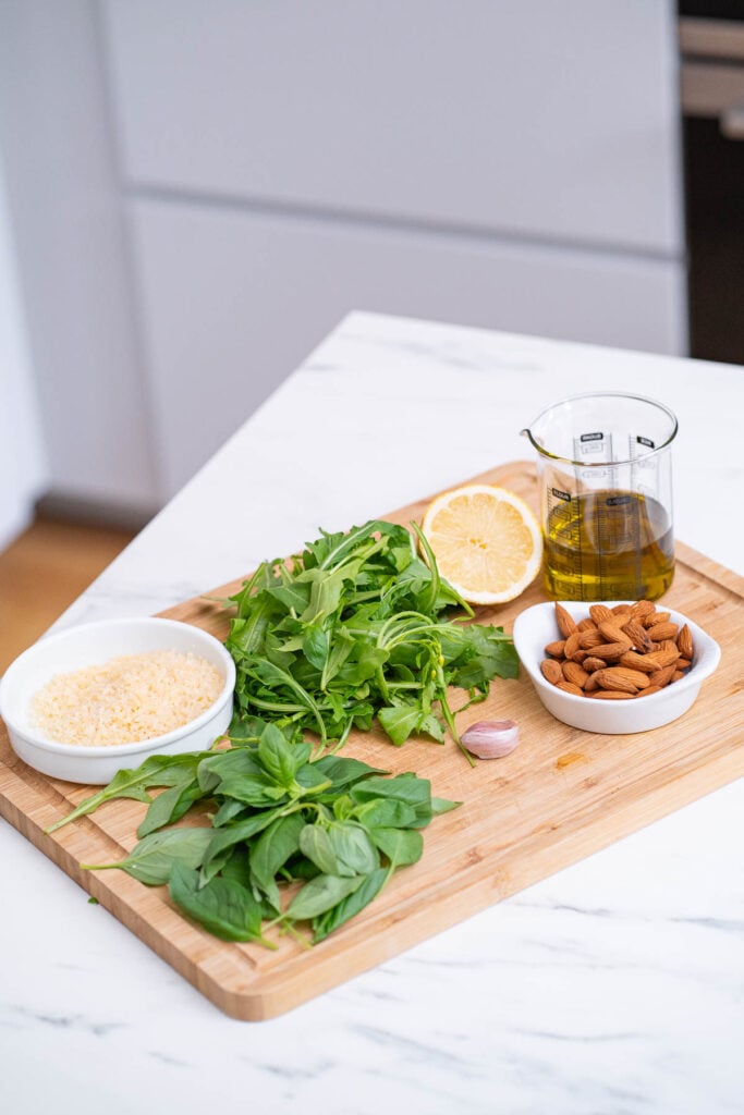 Une planche à découper avec du basilic frais, un quartier de citron, un petit bol de parmesan, des amandes entières, une gousse d'ail et un récipient en verre d'huile d'olive.