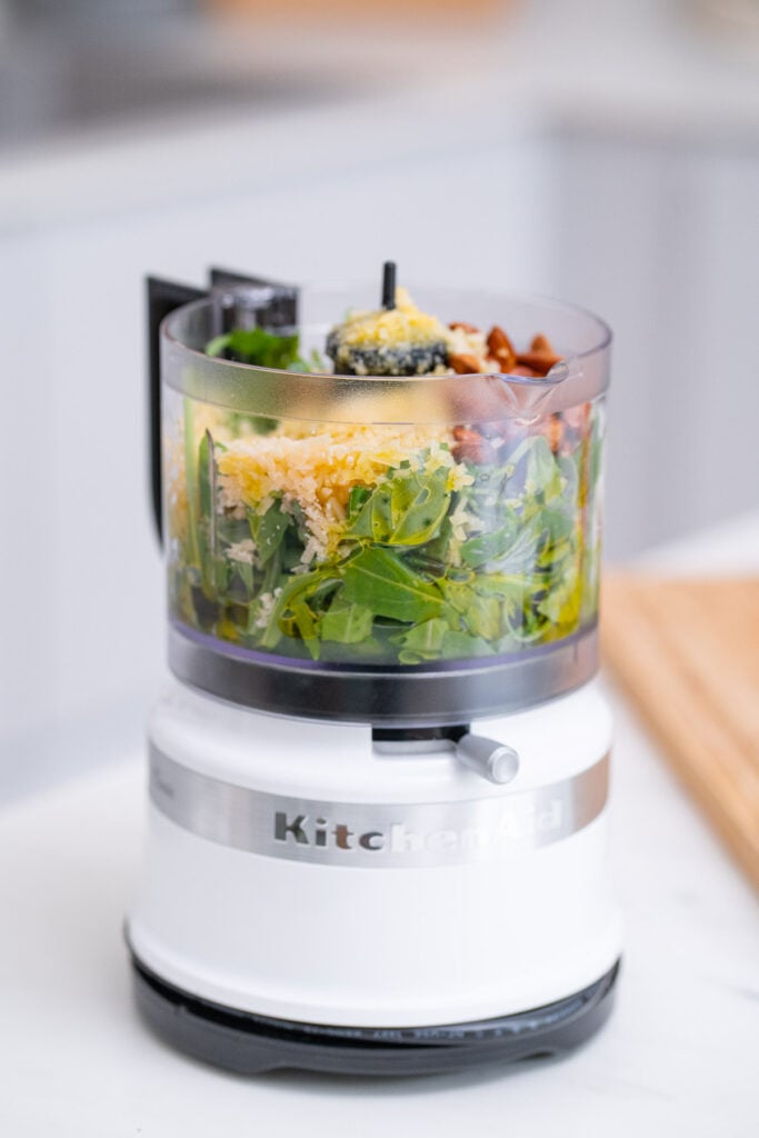 Un robot culinaire KitchenAid blanc est rempli d'ingrédients, notamment des légumes verts, du fromage râpé et des noix, placés sur un comptoir de cuisine.