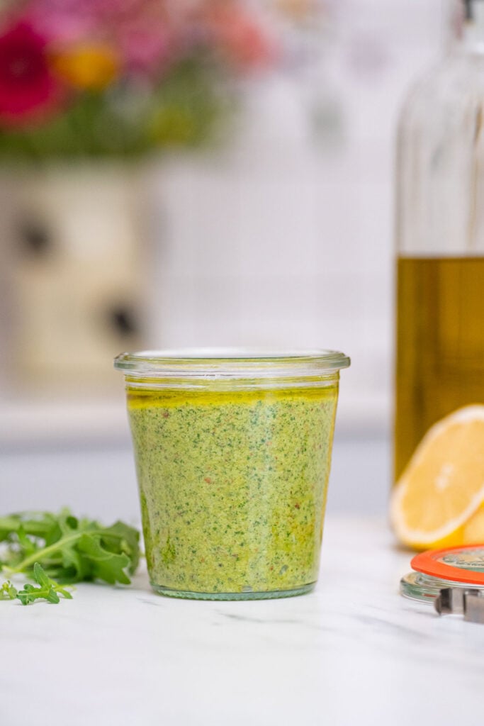 Un bocal en verre rempli de sauce pesto verte se trouve sur un comptoir de cuisine, entouré d'un demi-citron, d'herbes vertes fraîches et d'une bouteille d'huile.