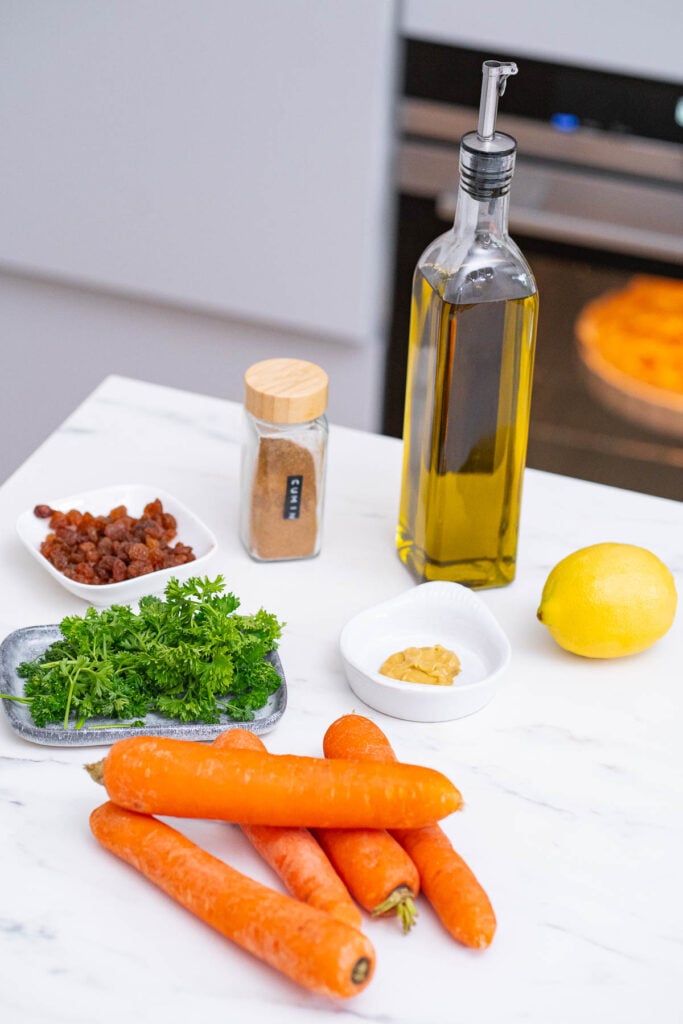 A bottle of olive oil, a lemon, ground cumin, golden raisins, fresh parsley, four carrots and a small dish of mustard laid out on a marble countertop.