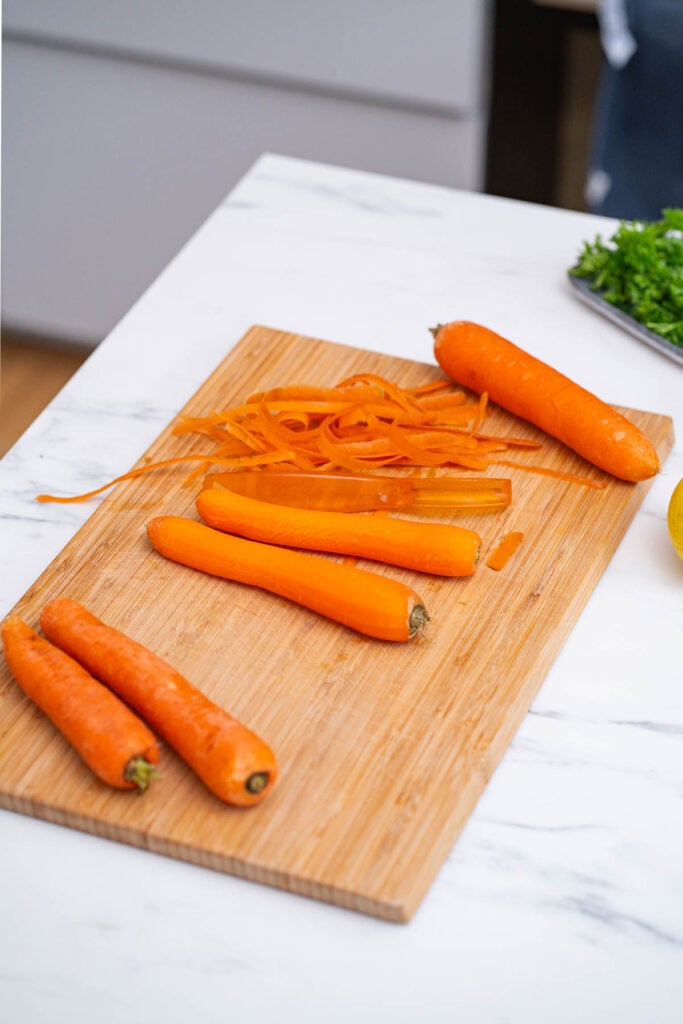 Four peeled carrots and carrot peels are on a wooden cutting board on a white marble countertop. A bouquet of parsley is in the background. 