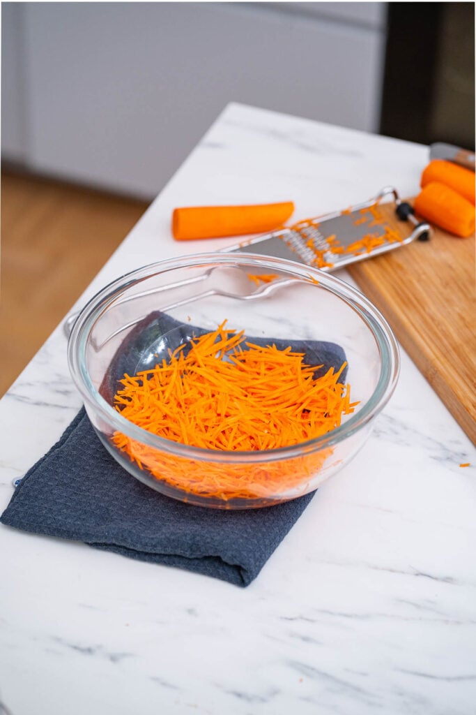 A glass bowl filled with grated carrots sits on a blue napkin on a marble countertop. A grater with additional carrot pieces is visible on a nearby wooden cutting board. 