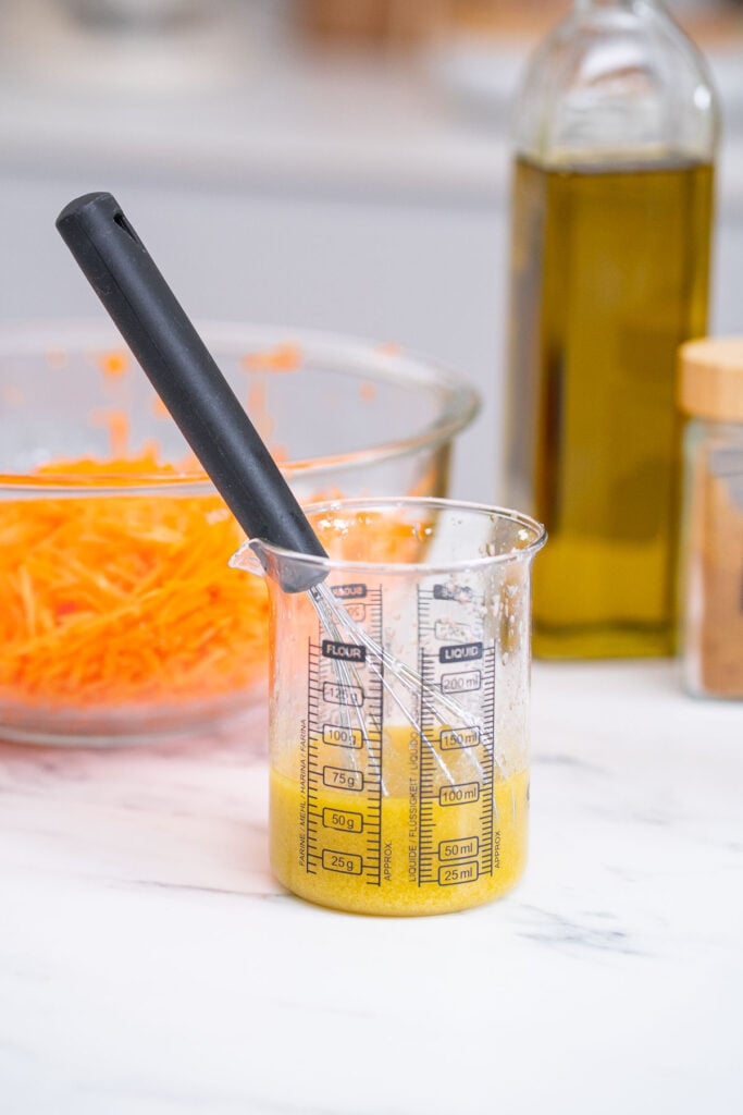 A small measuring glass filled with a leon and cumin vinaigrette and a whisk inside sits on a white countertop next to a bowl of grated carrots, a bottle of olive oil and a container with a wooden lid.