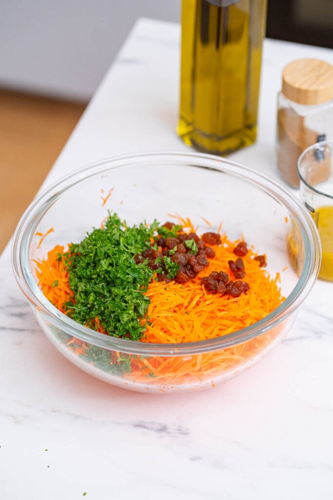 A clear glass bowl containing grated carrots, chopped parsley and raisins is placed on a marble countertop. Olive oil and spices are visible in the background. 