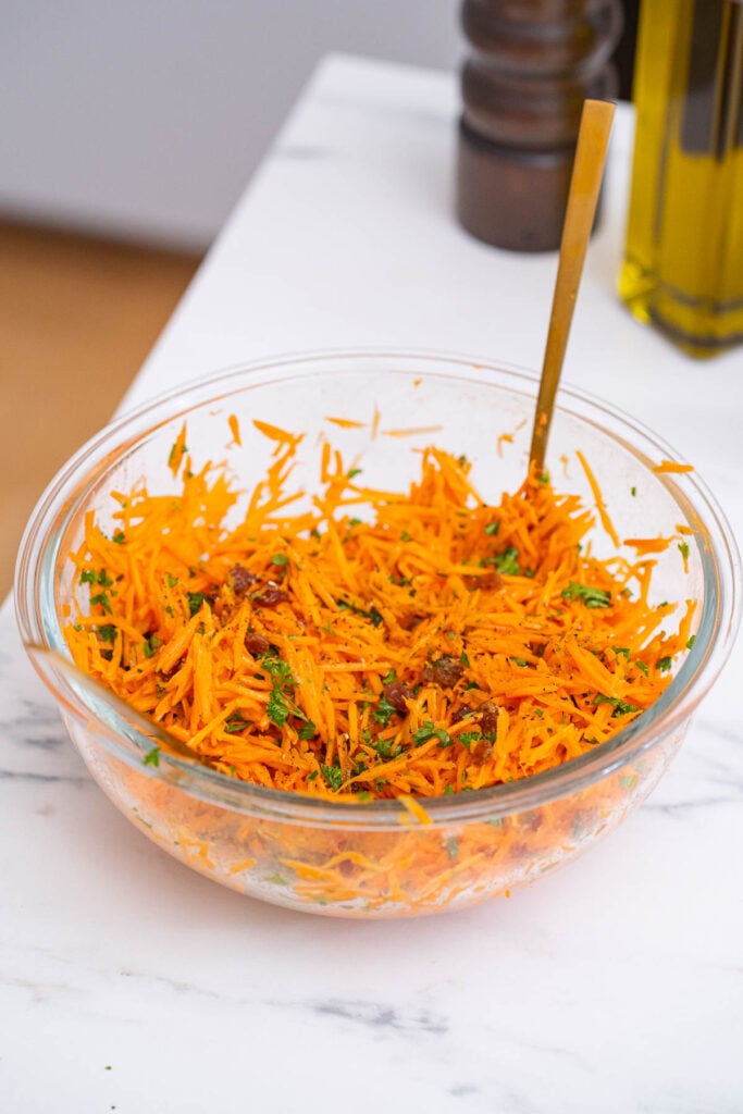 A glass bowl filled with shredded carrot salad mixed with herbs and raisins, set on a white marble countertop. A golden spoon is in the bowl. There are blurred objects in the background.  