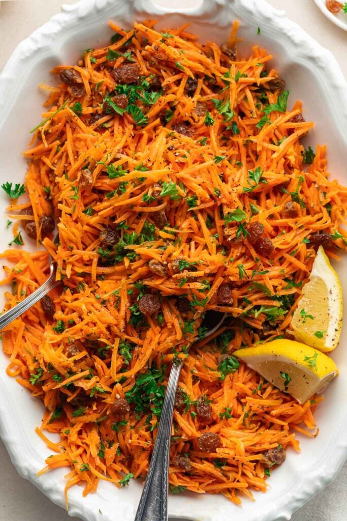 A white plate with a carrot salad garnished with parsley and lemon wedges. Two serving spoons are placed in the salad. 