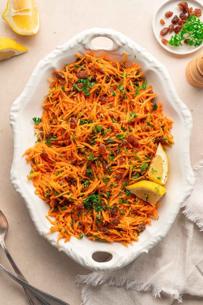 A white dish filled with shredded carrot salad garnished with parsley, raisins and two lemon wedges on the side. A piece of cloth, a slice of lemon and a spoon surround the dish. 