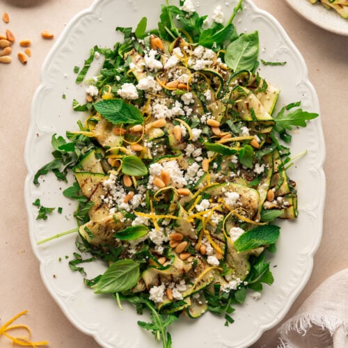 Un plateau blanc de salade de courgettes grillées garnie de fromage feta émietté, de pignons de pin, de menthe fraîche, de feuilles de basilic et de zeste de citron, placé sur une surface de couleur claire avec une serviette en tissu à proximité.