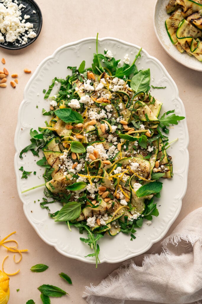 Un plateau blanc de salade de courgettes grillées garnie de fromage feta émietté, de pignons de pin, de menthe fraîche, de feuilles de basilic et de zeste de citron, placé sur une surface de couleur claire avec une serviette en tissu à proximité.